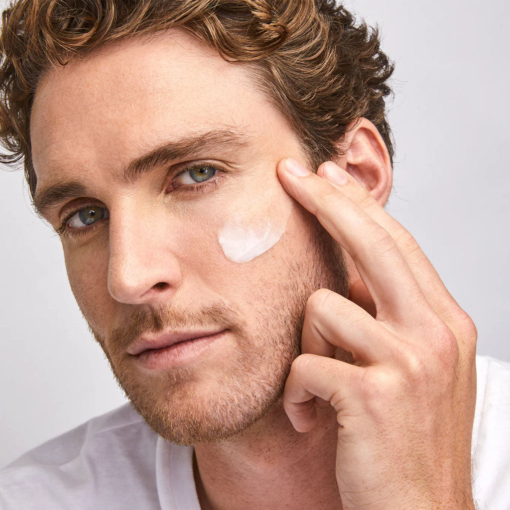 Man applying white Colorescience Barrier Pro Essential Moisturizer cream to his cheek against a light background.