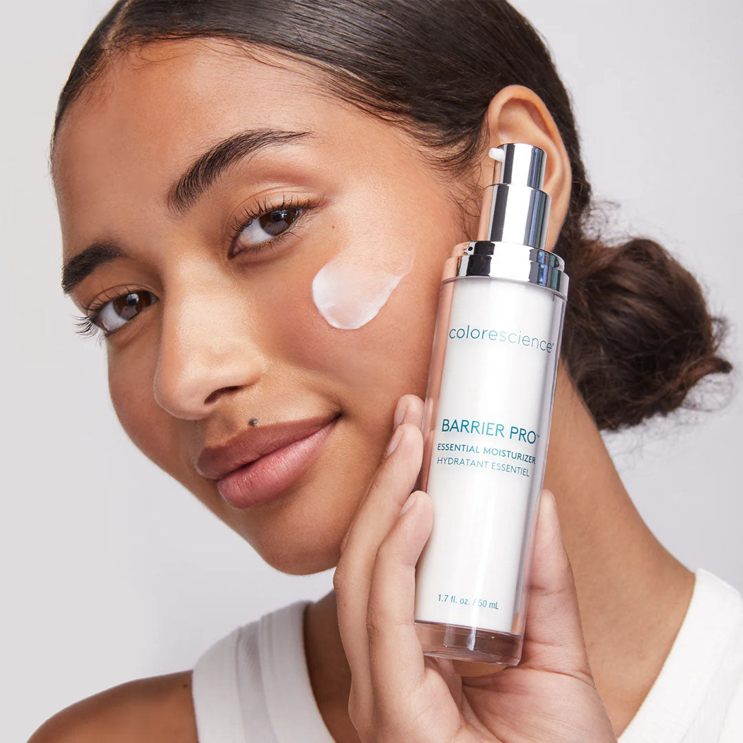 Woman holding a white 50ml Colorescience Barrier Pro Essential Moisturizer pump bottle with cream on her cheek.