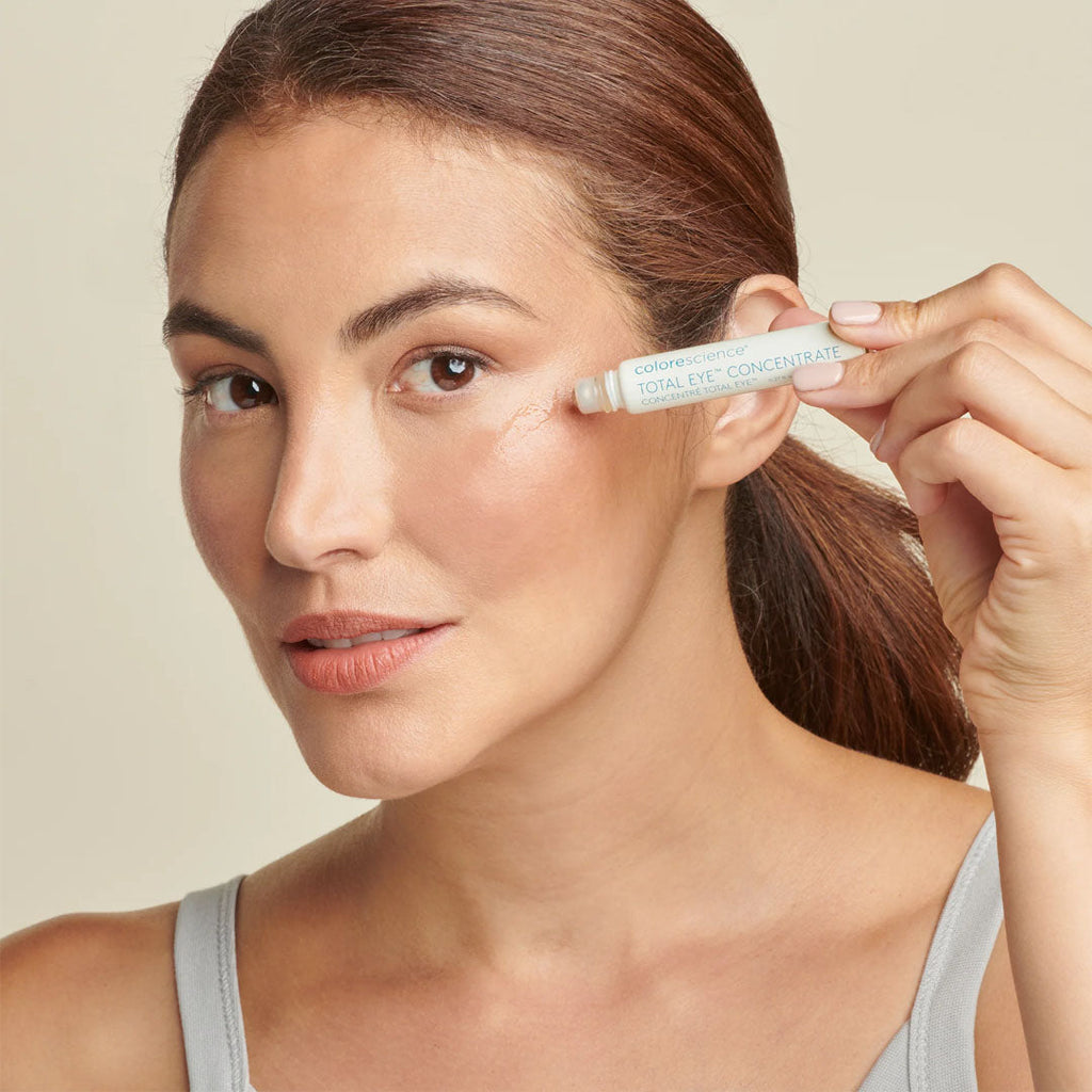 Woman applying white Total Eye Concentrate rollerball serum to her under-eye area for a refreshed, smooth appearance.