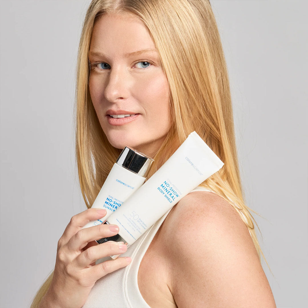 Blonde woman holding white Colorescience Total Protection SPF 50 sunscreen bottle and tube against a grey background.