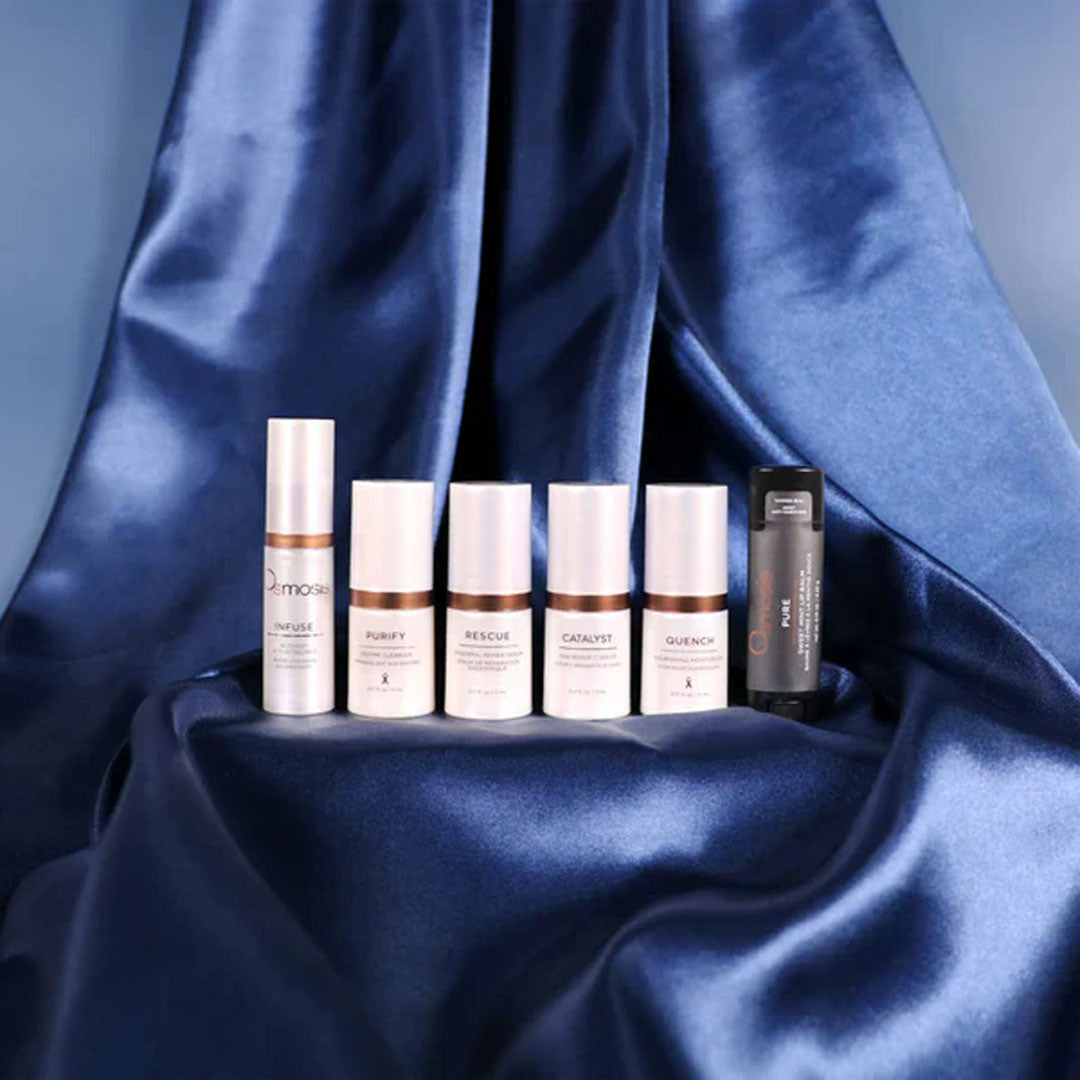 Five white and one black travel-sized skincare bottles on a draped navy blue satin background.