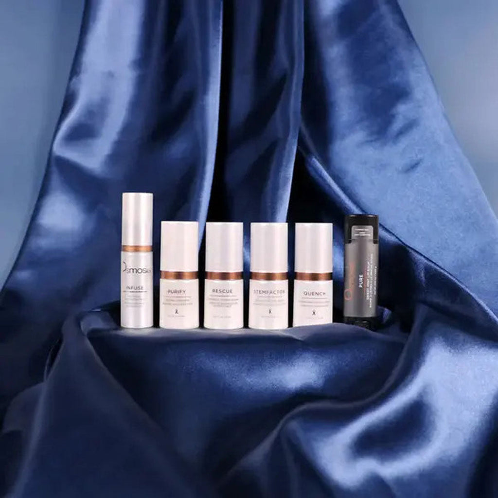 Five white and one dark gray mini skincare bottles on a draped blue satin fabric.
