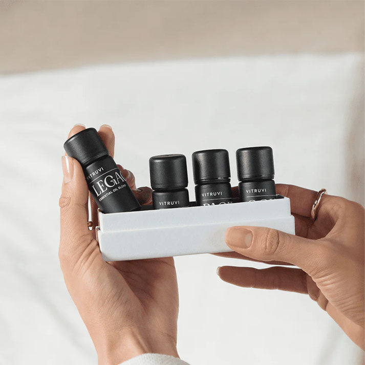 Hands holding a white tray with four small black Vitruvi essential oil bottles, one labeled "Legacy.