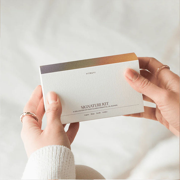 Hands holding a white Vitruvi Signature Kit box with a colorful gradient top and essential oil blends.